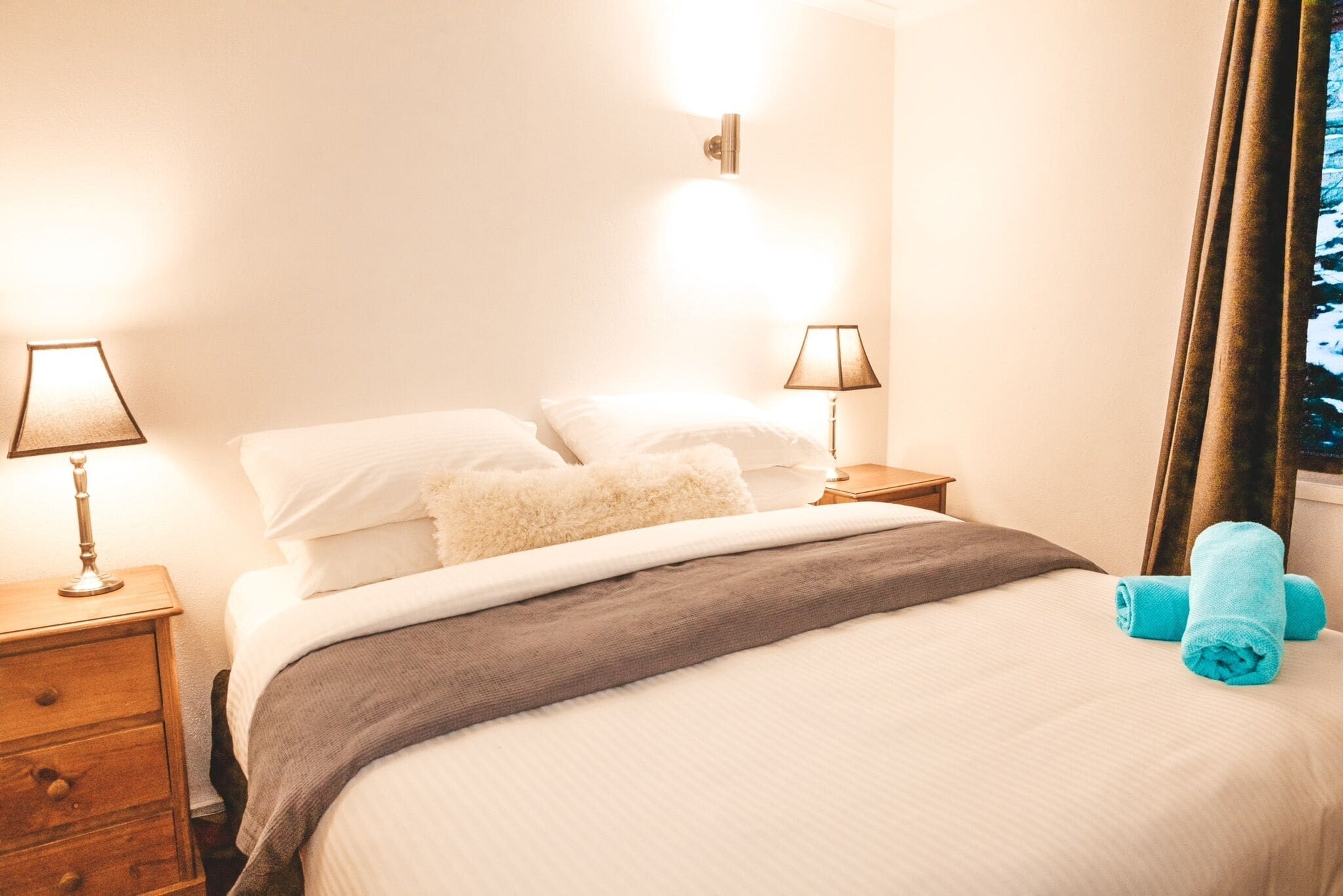 A neatly made bed with white linens and a gray blanket, two bedside lamps, a wooden nightstand, and a window with curtains.