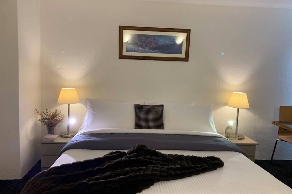 A hotel room with a bed, two bedside lamps, a framed picture on the wall, and a small table with a vase.