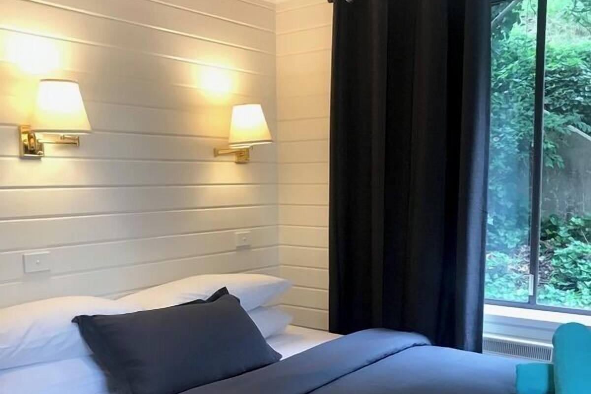 A hotel room with a bed, two wall-mounted lamps, a window with curtains, and a view of greenery outside.