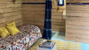 A room with wooden walls, a bed with floral bedding, a ceiling fan, a blue curtain, a small table with magazines, and a coat rack. - Charming house 3 kms from Caen.  Comfort, light, location -> Coup de coeur! (Fleury-sur-Orne)