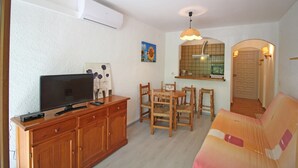 A living room with a flat-screen TV, wooden furniture, a dining table with chairs, a sofa, and a kitchen area with a tiled backsplash and a hanging light fixture. - Studio in Empuriabrava near Sandy Beach (Empuriabrava)