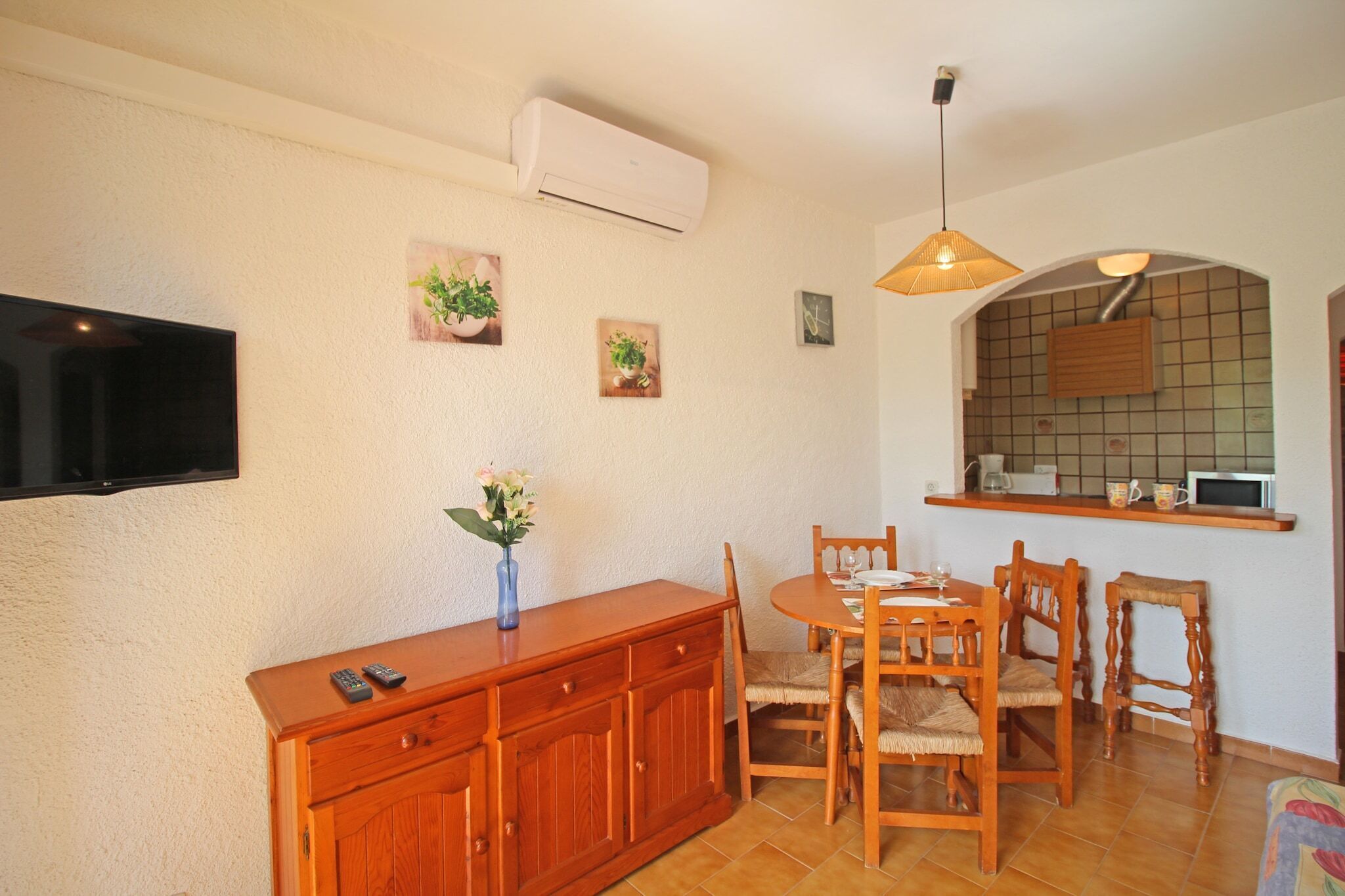 Een eethoek met een houten tafel en stoelen, een aan de muur bevestigde televisie, en een keuken met een magnetron en een plank boven het aanrecht.
