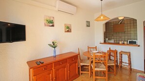 Ein Essbereich mit Holztisch und -stühlen, einem an der Wand montierten Fernseher sowie einer Küchenzeile mit Mikrowelle und einem Regal über der Arbeitsfläche.