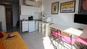 A compact, well-lit room with a kitchenette, a dining area with a table and chairs, and a bed with a yellow bedspread and a straw hat. - Studio in Empuriabrava with Sea Views (Empuriabrava)