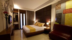 Une chambre d’hôtel avec un grand lit, une tête de lit en bois, un canapé rouge, une table de chevet, un miroir et un mur orné d’une fresque représentant un paysage urbain.