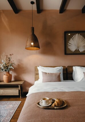A cozy bedroom with a wooden headboard, a hanging pendant light, a bedside table with a vase of dried flowers, and a tray with croissants and a cup of coffee.