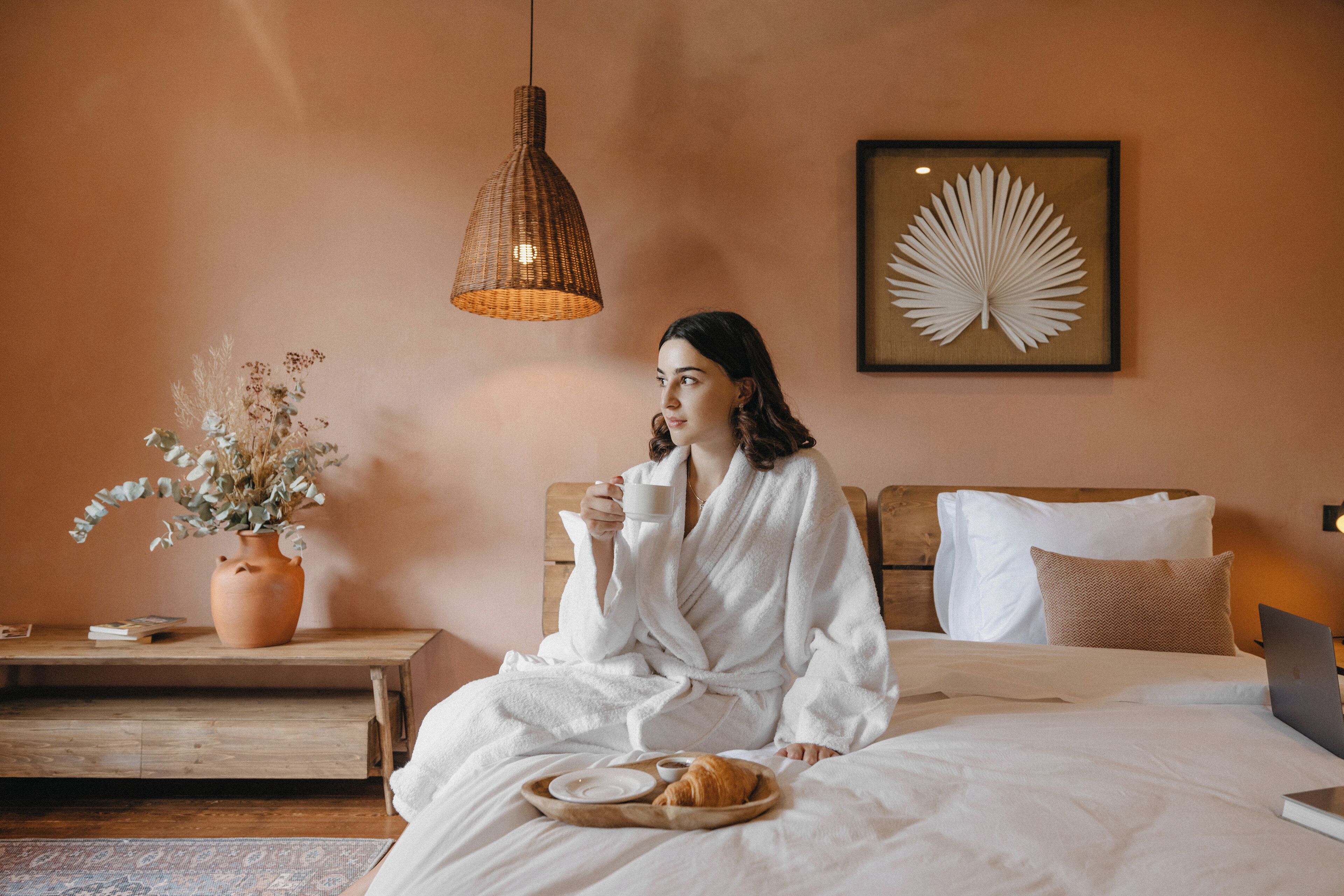 Eine Person in einem weißen Bademantel sitzt auf einem Bett, neben ihr ein Laptop, ein Tablett mit einem Croissant und einer Tasse.
