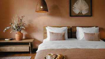 Una cama bien hecha con almohadas, una mesita de noche con un jarrón de flores secas, una obra de arte enmarcada en la pared y una bandeja con cruasanes y café sobre la cama.