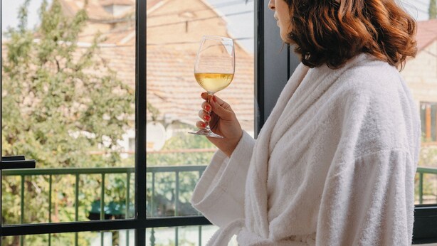 A person in a white bathrobe holding a glass of white wine, standing by a large window with a view of buildings and greenery. - Hameau Mukhrani (Mtskheta)