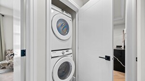 A laundry room with two stacked washing machines, a door leading to a bathroom, and a patterned chair in the background.