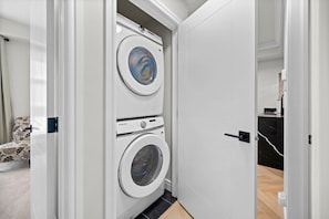 Uma lavanderia com duas máquinas de lavar empilhadas, uma porta que dá para um banheiro e uma cadeira estampada ao fundo.