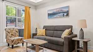 A living room with a grey sofa, two side tables with lamps, a patterned armchair, a coffee table, and a large window with a yellow curtain.