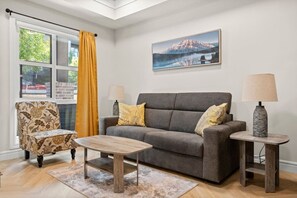 Uma sala de estar com um sofá cinzento, duas mesas laterais com luminárias, uma poltrona estampada, uma mesa de centro e uma janela grande com cortina amarela.