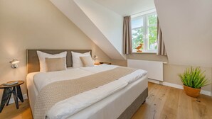 A bedroom with a large bed, a bedside table, a lamp, a window with curtains, and a potted plant.