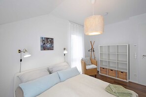 Un dormitorio con una cama, dos lámparas de noche, una silla de mimbre, un armario y un cuadro en la pared.