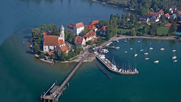 Vue aérienne d’une ville au bord d’un lac, avec une église, des immeubles résidentiels et un port de plaisance où sont amarrés des bateaux.