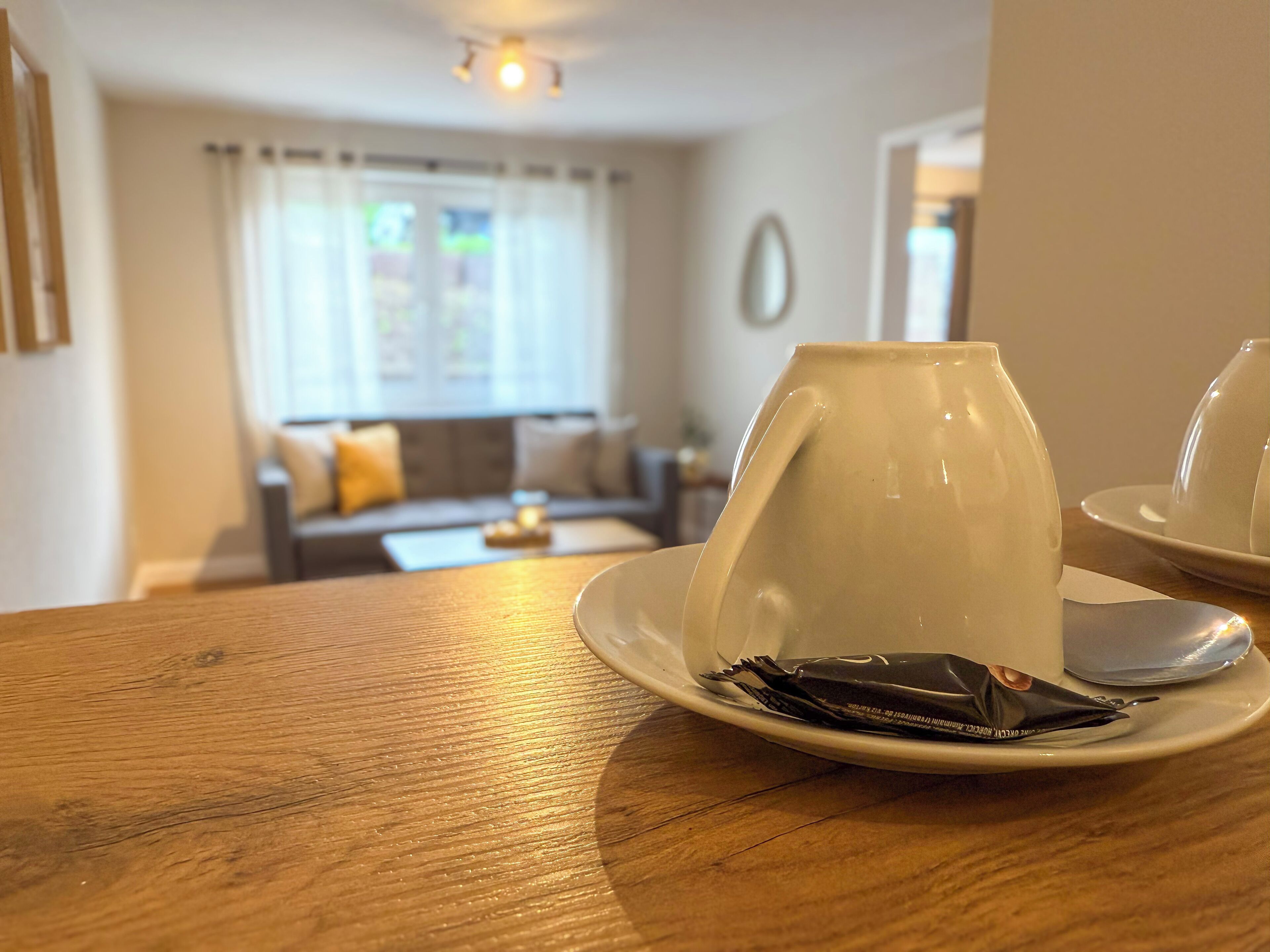 A dining table with a white cup and saucer, a spoon, and a chocolate bar. In the background, there is a living room with a sofa, a coffee table, and a window with curtains.