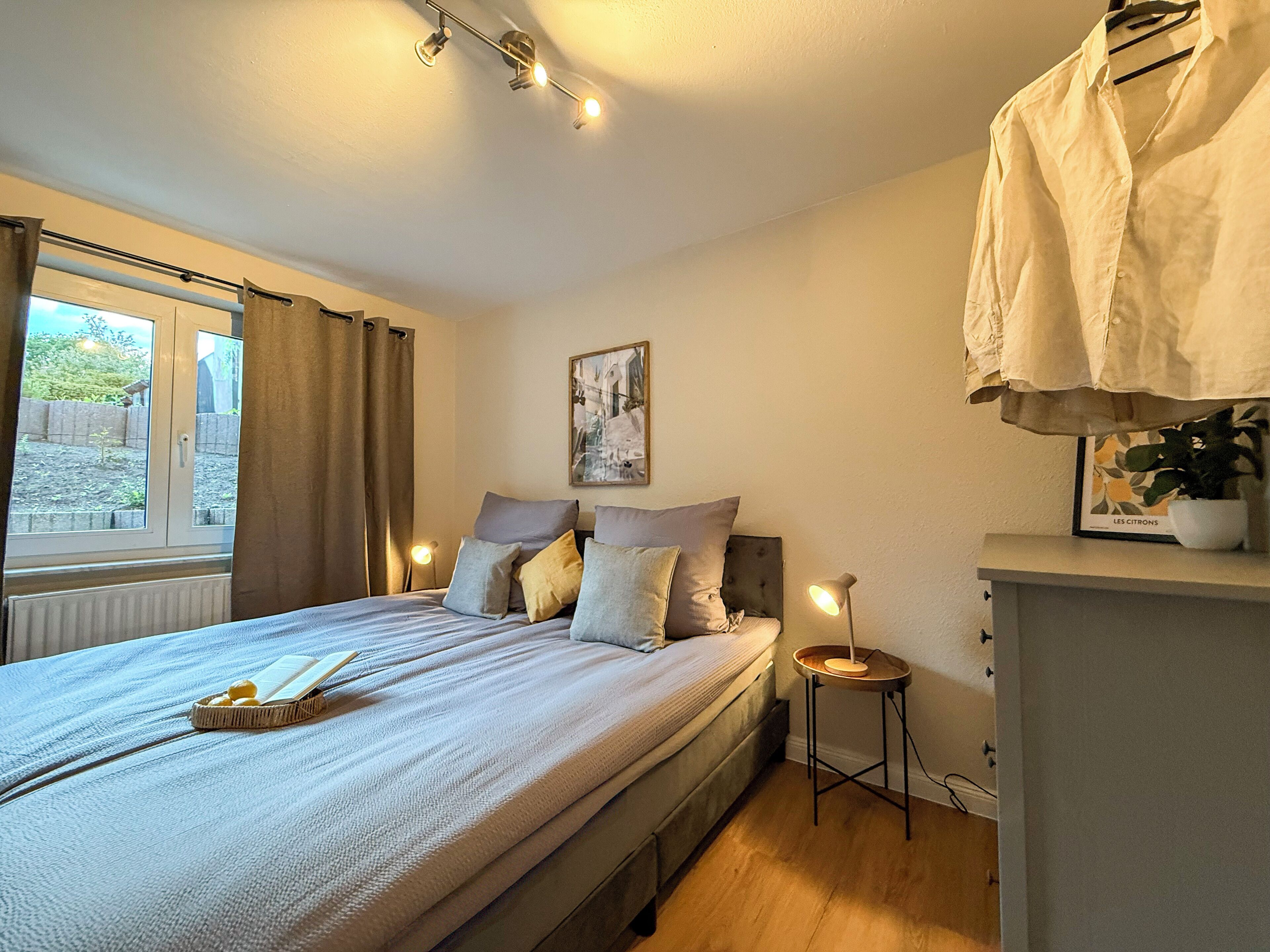 A bedroom with a bed, bedside lamps, a window with curtains, a bedside table, and a wall-mounted shelf with a plant.