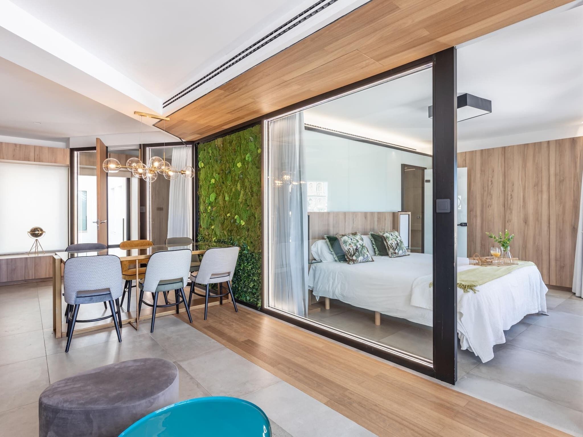 Une chambre d’hôtel moderne dotée d’une grande porte-fenêtre coulissante donnant sur un balcon, d’un lit avec une couette blanche, d’une petite table avec un vase et d’un coin salon avec des chaises et une petite table.
