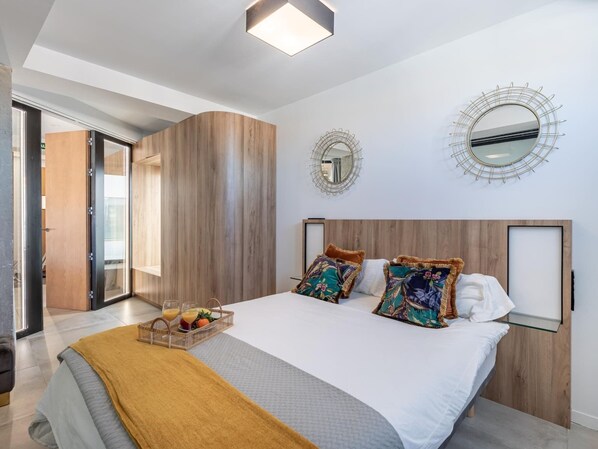 Une chambre à coucher moderne avec un grand lit, une tête de lit en bois, deux miroirs muraux et une table de chevet en verre.