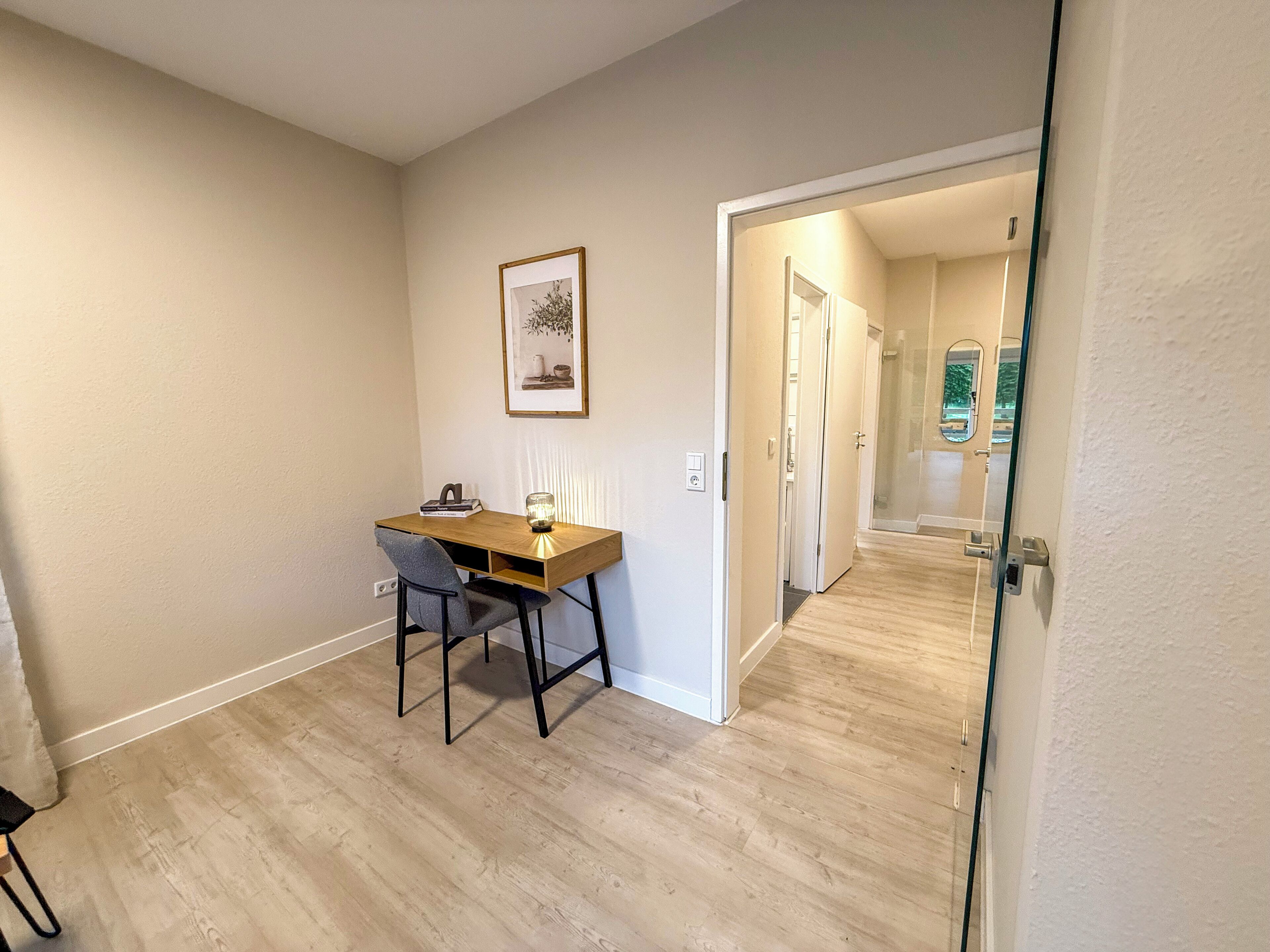 Une petite pièce bien éclairée, avec un bureau en bois, une chaise et un tableau encadré accroché au mur.