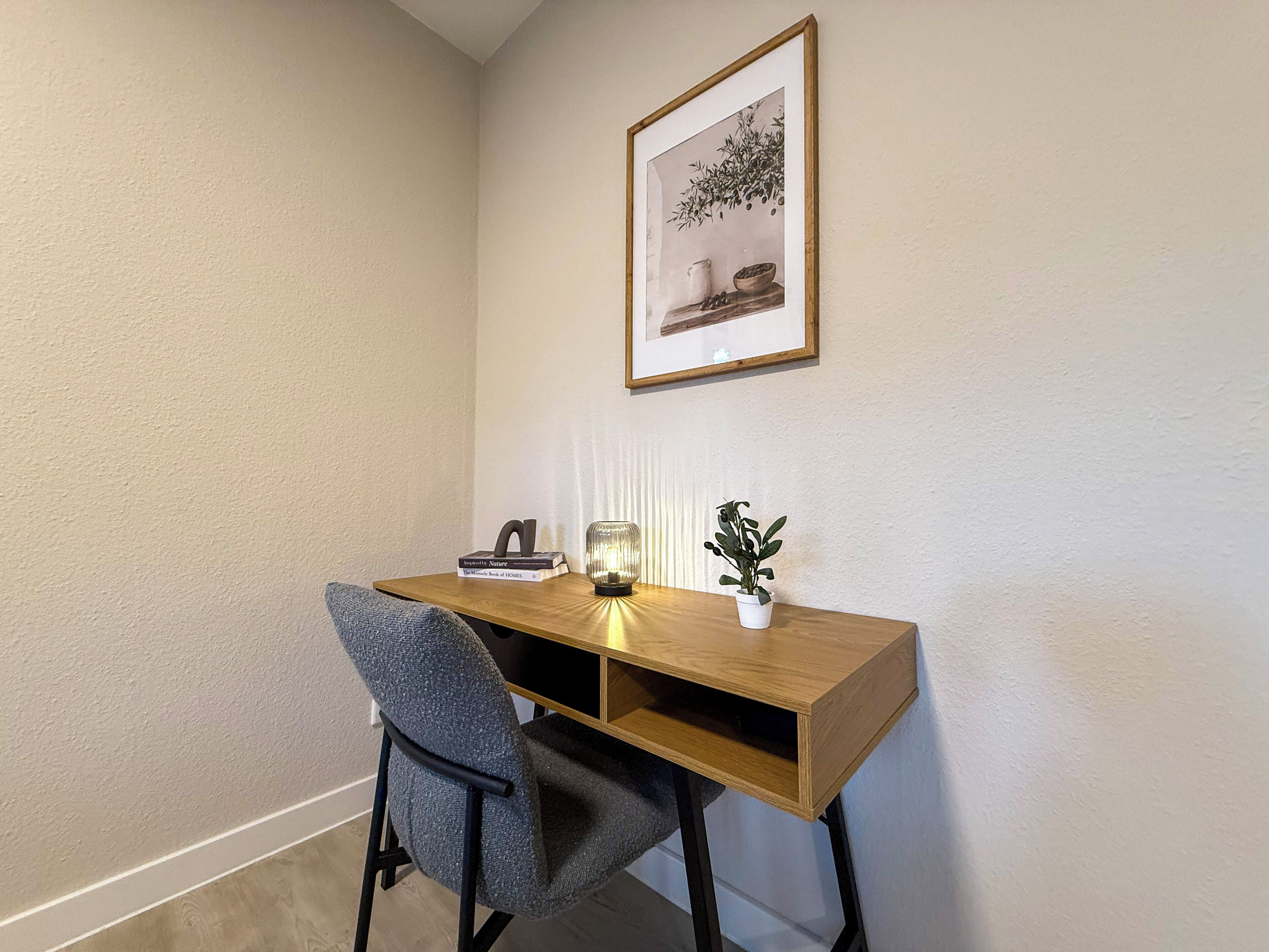 A corner office with a wooden desk, a chair, a framed picture on the wall, a small potted plant, and a lamp.