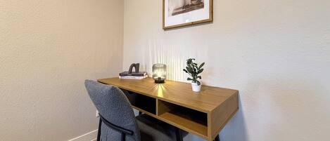 A corner office with a wooden desk, a chair, a framed picture on the wall, a small potted plant, and a lamp.