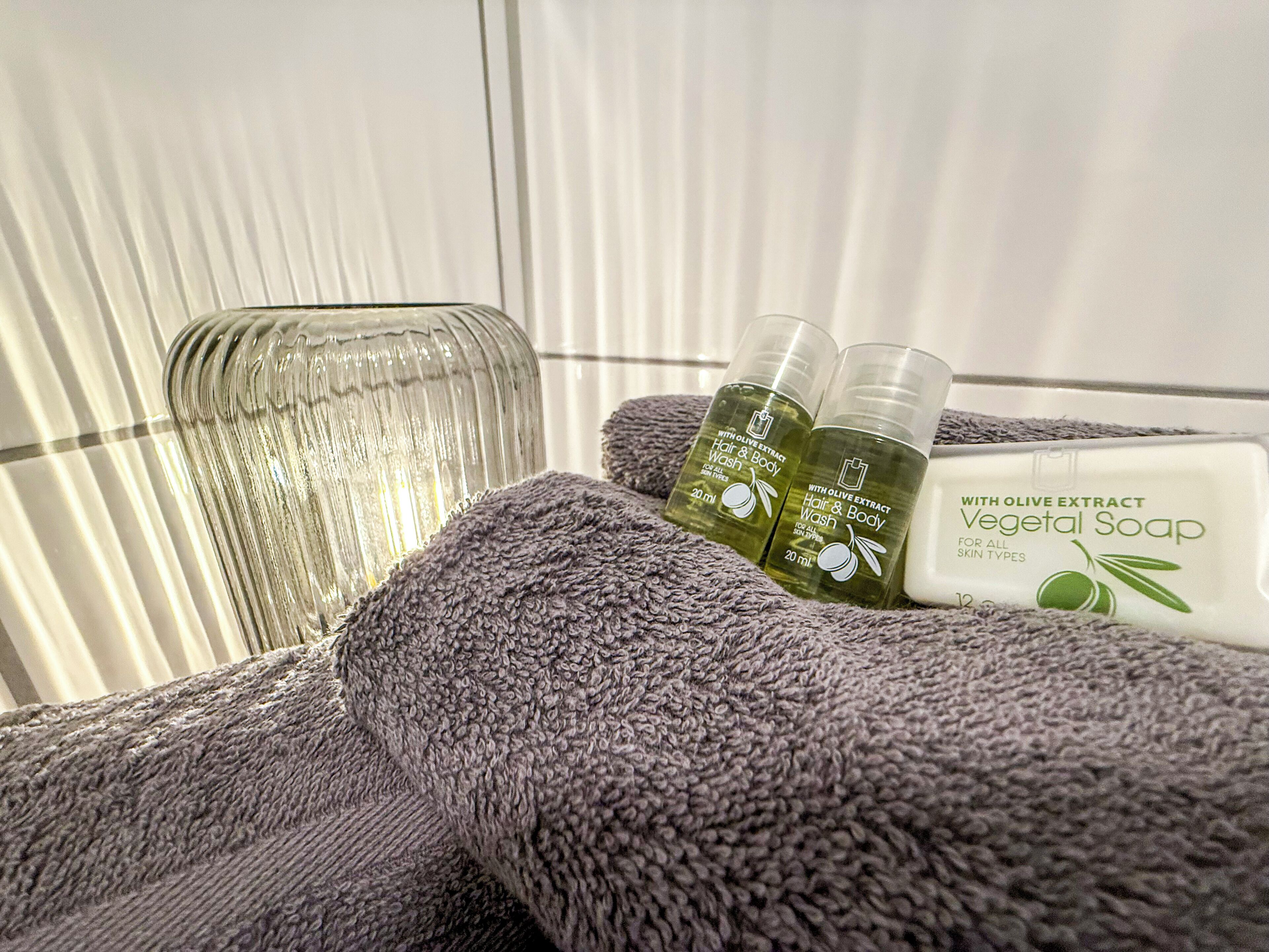 Une salle de bain avec une douche à parois de verre, une serviette violette et des flacons de produits pour les cheveux et le corps à base d’extrait d’olive.