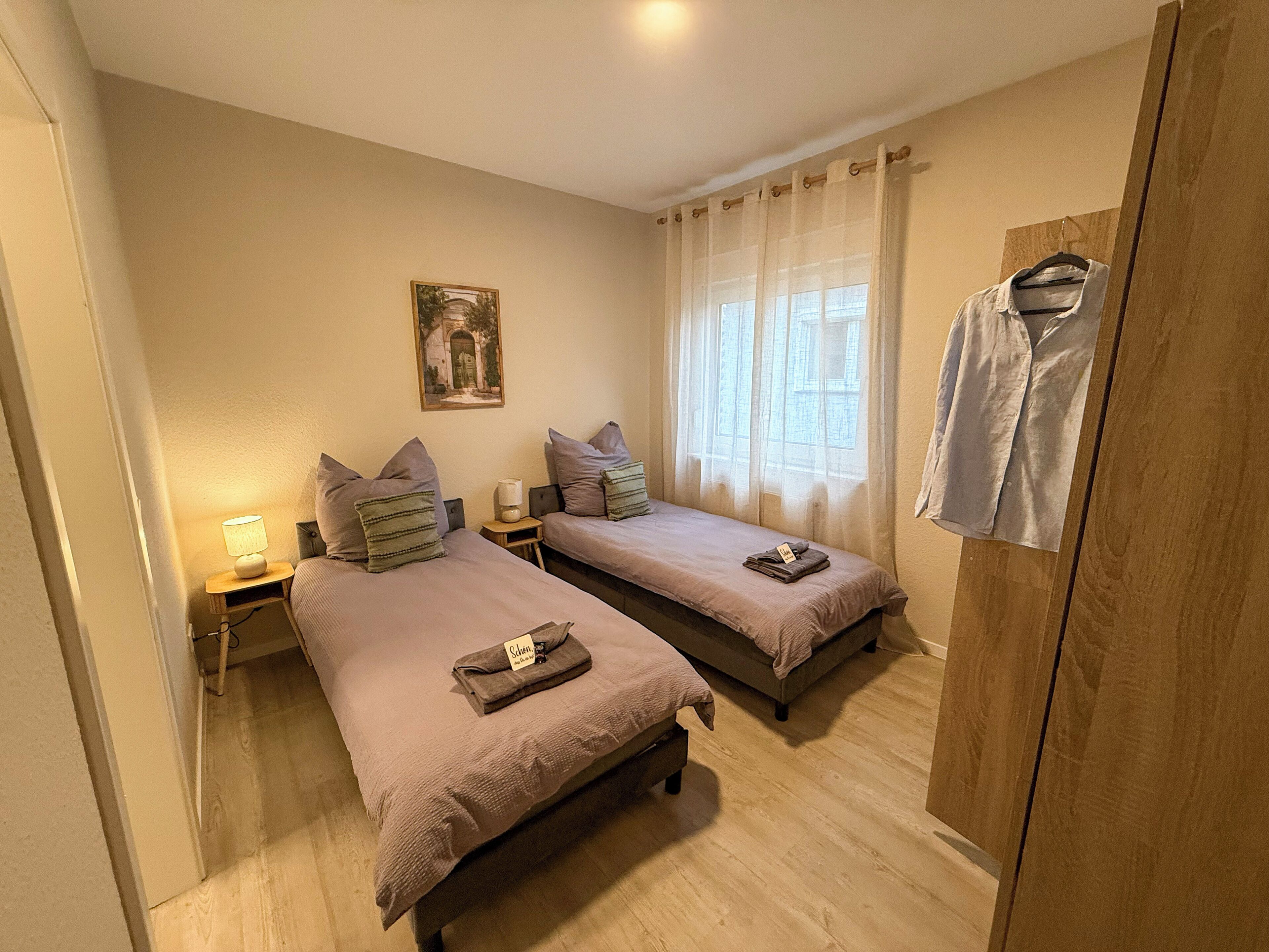 Ein Schlafzimmer mit zwei Einzelbetten, einem Holzschrank, einem Nachttisch mit Lampe und einem Fenster mit durchsichtigen Vorhängen.