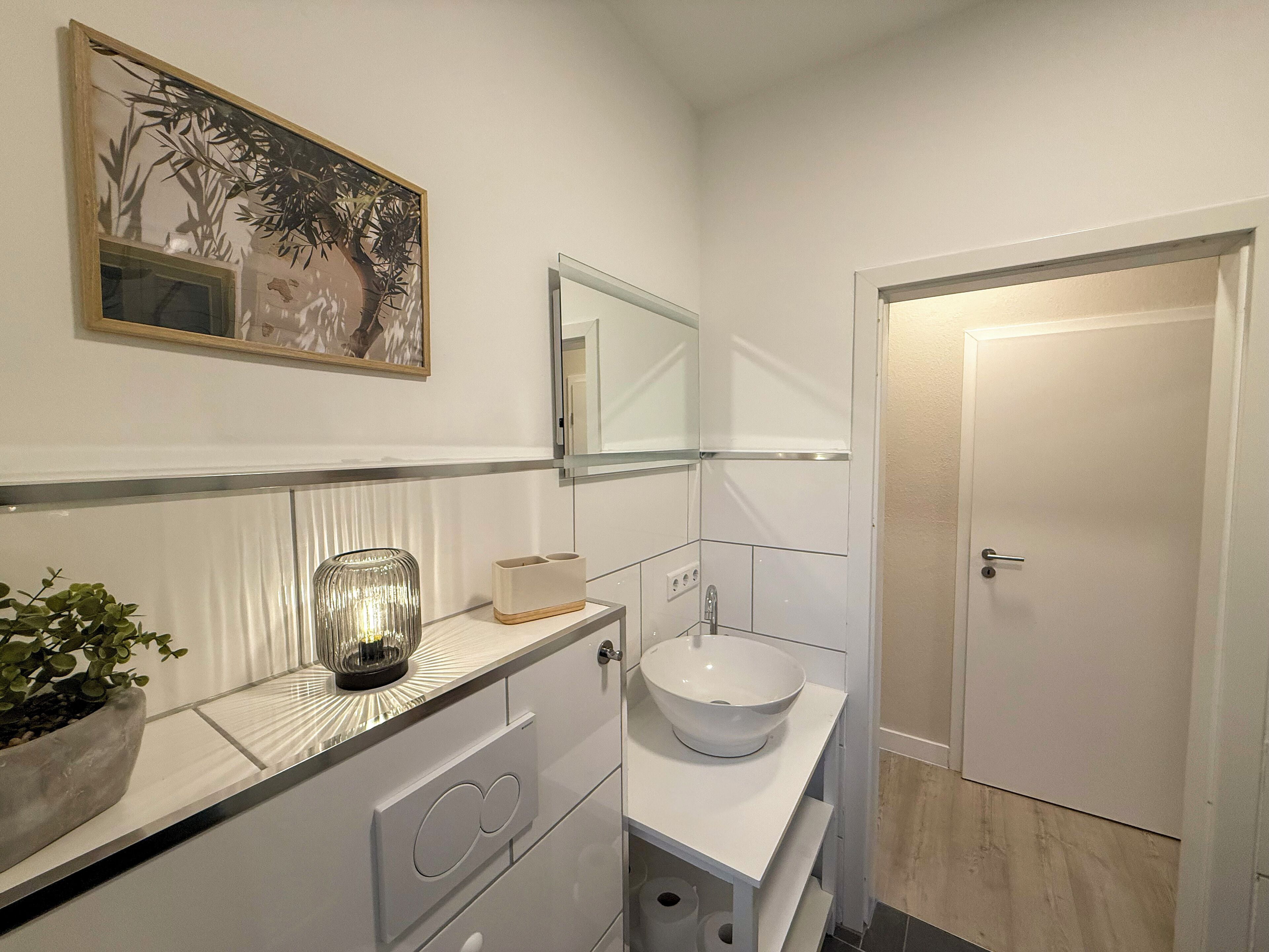Une salle de bain moderne avec un lavabo blanc, un miroir et une plante en pot posée sur le meuble-lavabo.