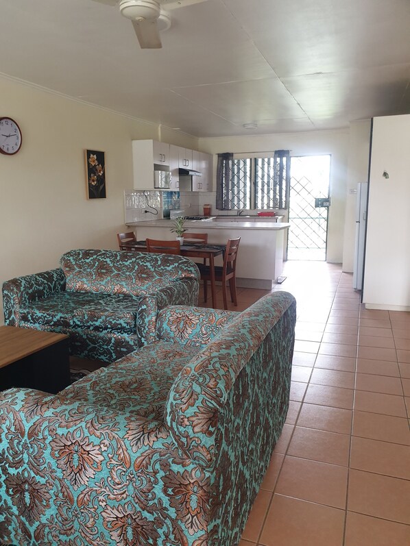 Una sala de estar con sofás estampados, un comedor, una cocina con armarios blancos y vistas a través de una ventana con reja de metal.
