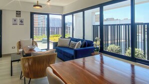 A modern living room with a blue sofa, a wooden dining table, and large windows offering a view of the cityscape. - w* | Well-Located 2BR in Miraflores (Miraflores)