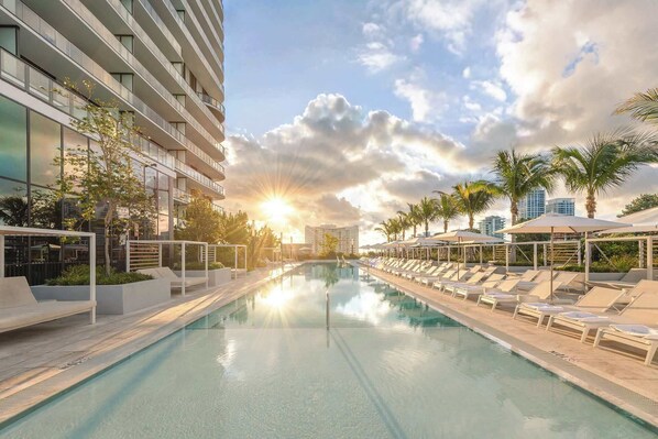 A modern hotel with a swimming pool, sun loungers, and palm trees.