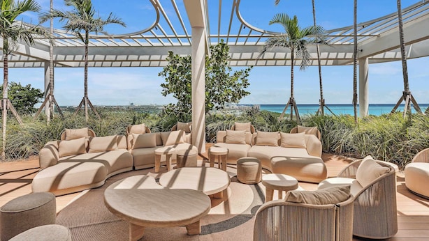 Un espace de détente extérieur doté d’un canapé incurvé, de tables rondes et de fauteuils en osier, offrant une vue sur la côte.