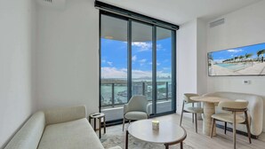 Un salon moderne avec un canapé, une table ronde et des chaises, une grande porte-fenêtre coulissante donnant sur un balcon, et un tableau accroché au mur.