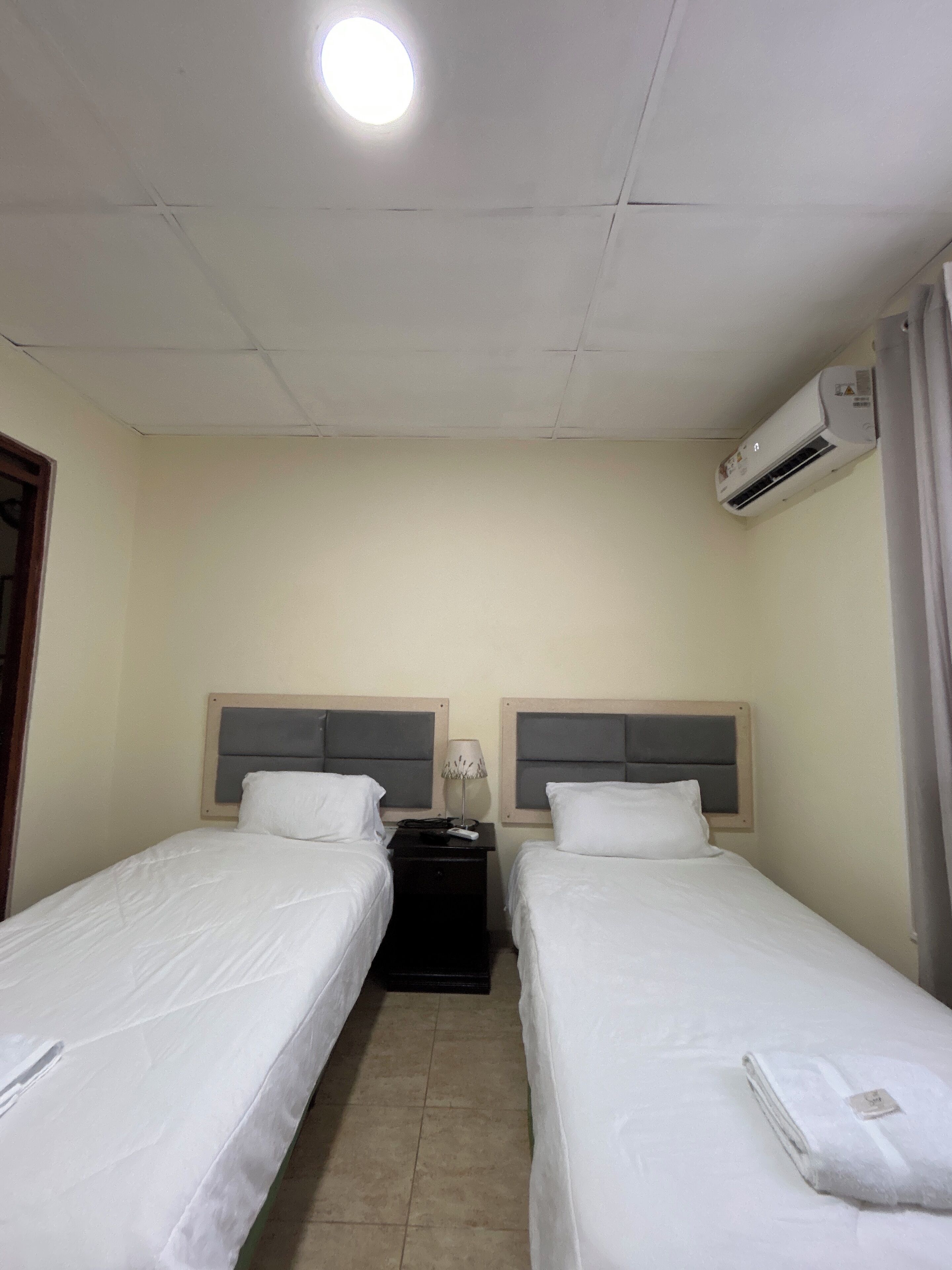 A hotel room with two single beds, a nightstand, an air conditioning unit, and a ceiling light.