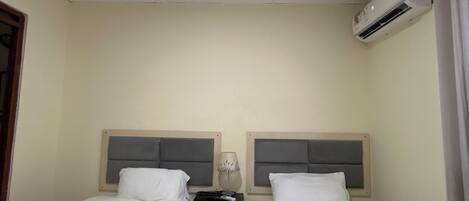 A hotel room with two single beds, a nightstand, an air conditioning unit, and a ceiling light.