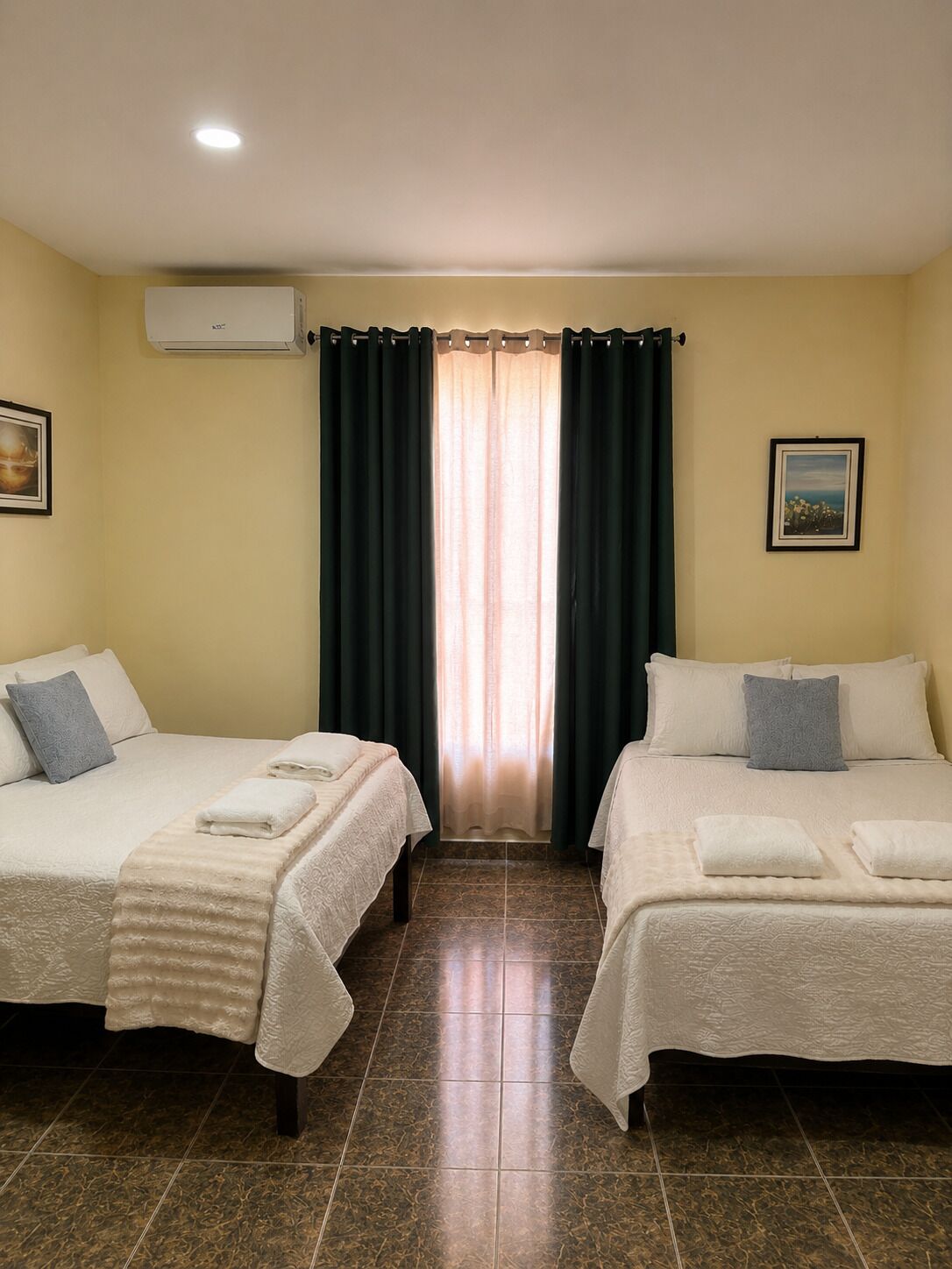 A hotel room with two beds, a window with curtains, a picture on the wall, and an air conditioning unit.