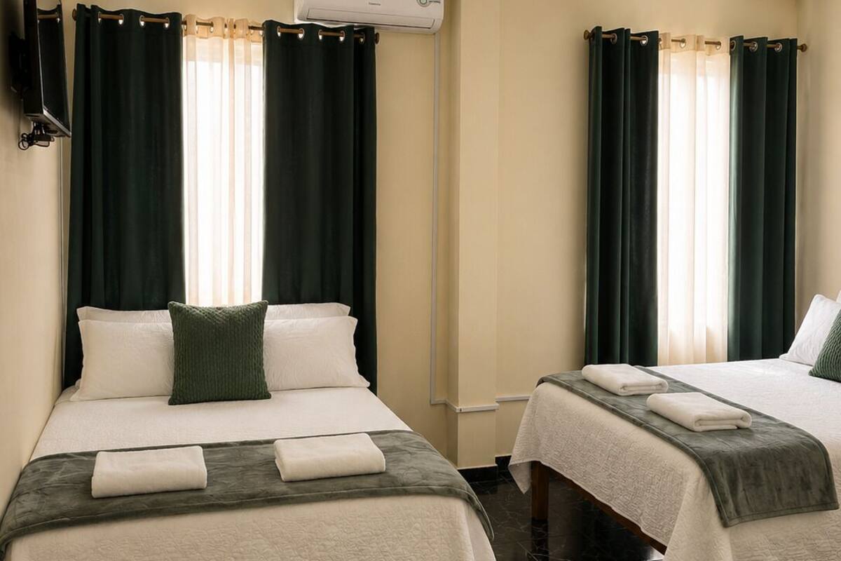 Two beds with dark green curtains, a flat-screen TV, and an air conditioning unit.