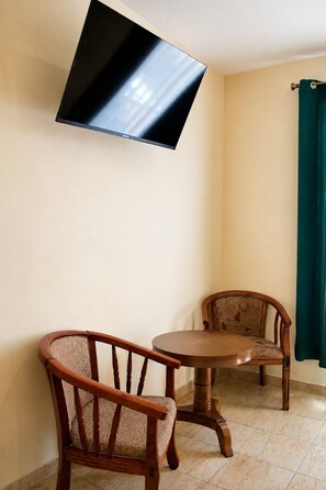 A room with a flat-screen TV mounted on the wall, two wooden chairs with cushioned seats, a round wooden table, and a green curtain on the window. - Hotel Crown Mirage (David Sur)