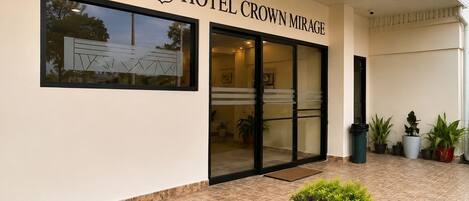 A hotel entrance with a large glass door, a sign reading 'HOTEL CROWN MIRAGE,' and potted plants in the foreground.