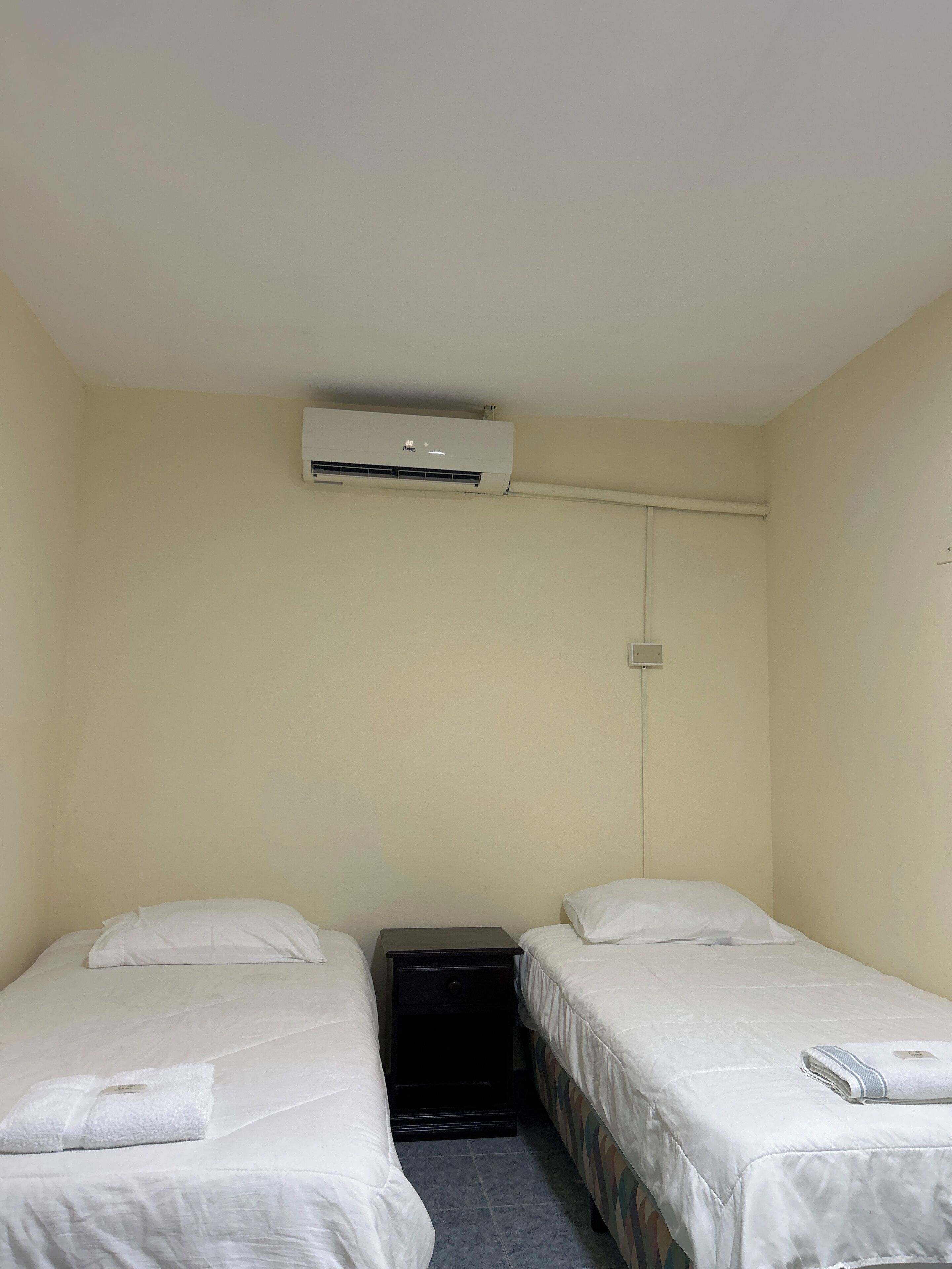 Een kleine, eenvoudige verblijfplaats met twee bedden, een nachtkastje en een airconditioning aan de muur.