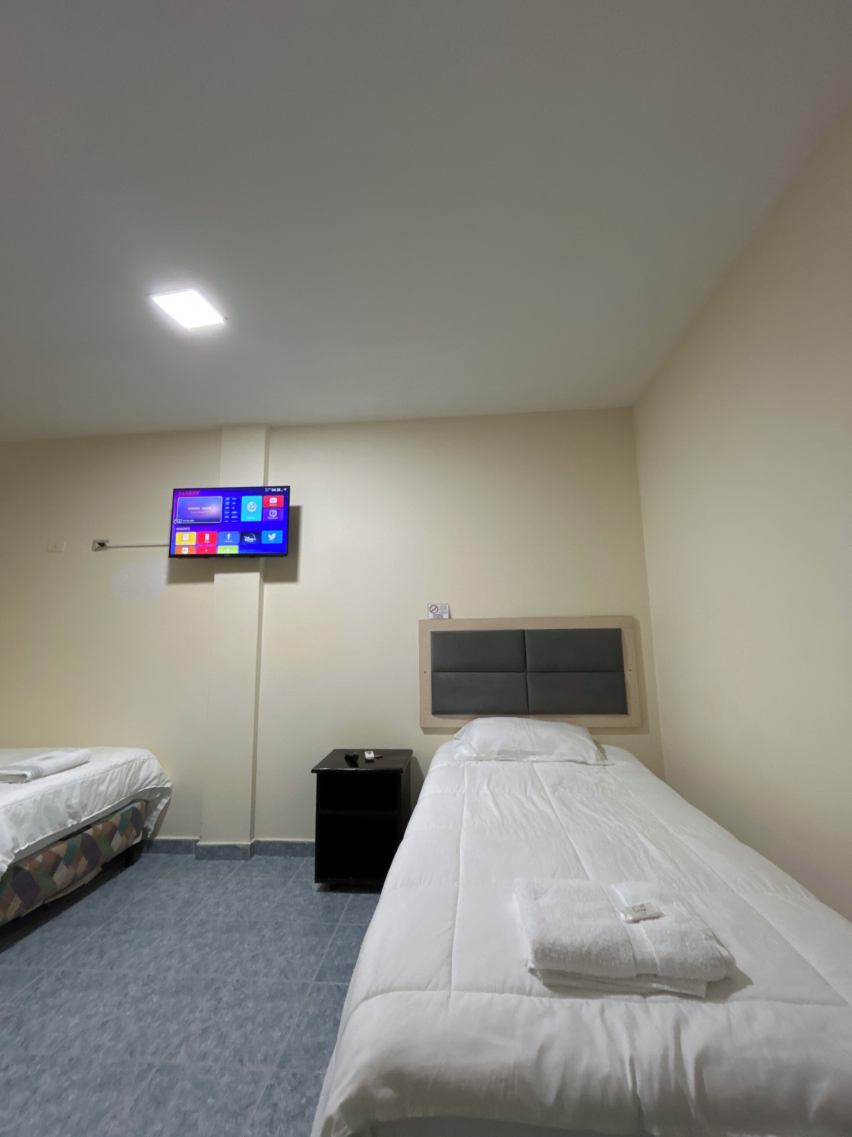 A hotel room with a single bed, a small bedside table, a TV mounted on the wall, and a plain wall with a sign.