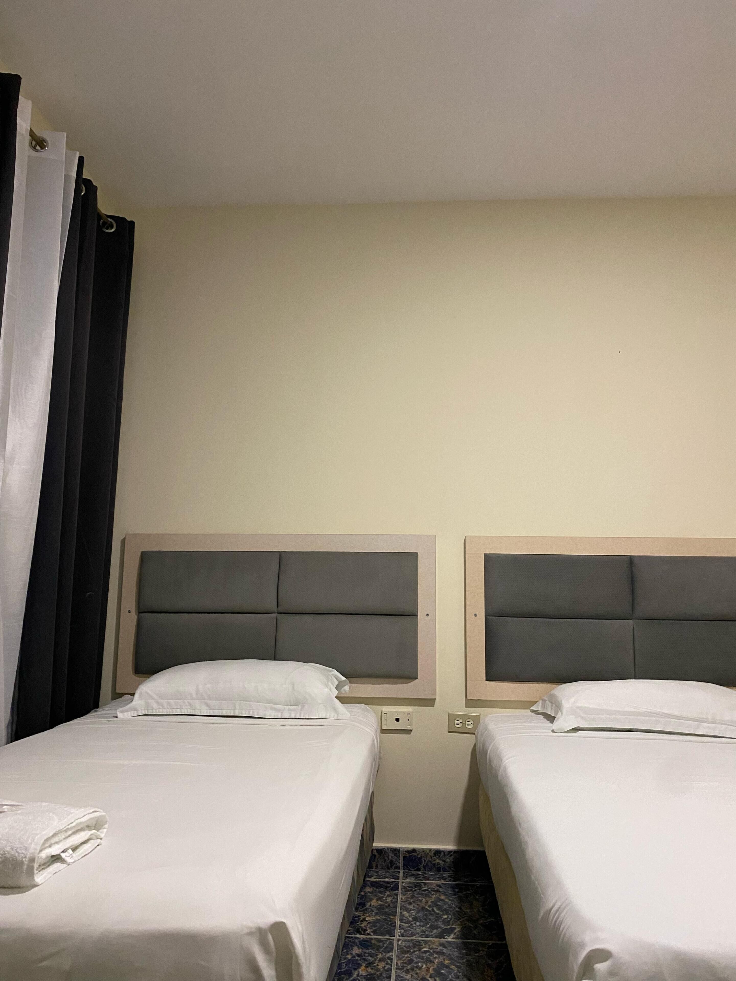 A hotel room with two beds, each with a headboard, a white towel on the left bed, and a power outlet on the wall.