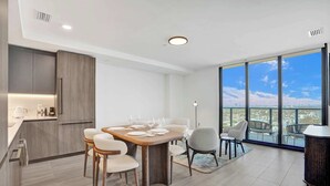 Ein zeitgemäßer Essbereich mit Holztisch, gedeckt für vier Personen, eine Küche mit integrierten Geräten und ein großes Fenster mit Blick auf die Stadt.