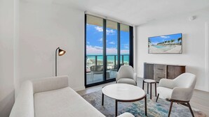 Ein modernes Wohnzimmer mit großem Fenster, einer Couch, einem Couchtisch, einem Sessel und einem flachbildfernseher an der Wand.