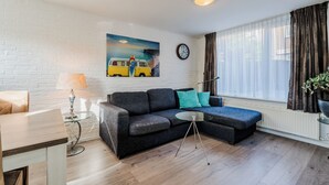 Una sala de estar moderna con un sofá modular, una pequeña mesa de vidrio, una lámpara, un reloj y un ventanal grande con cortinas.