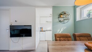 Una sala de estar moderna con un televisor de pantalla plana montado en la pared, una mesa de comedor con sillas y una cocina al fondo.