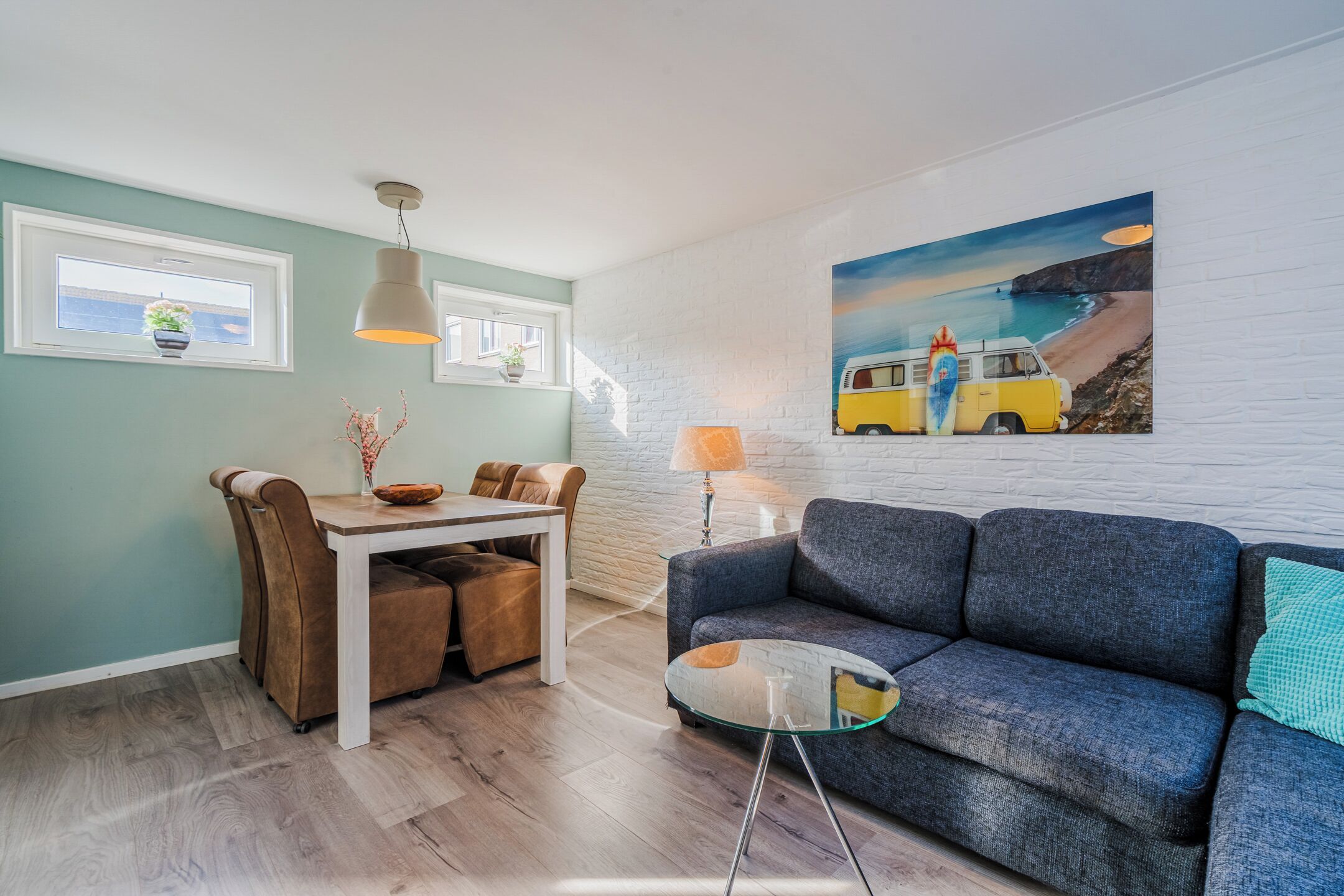 Een moderne woonkamer met een grijze bank, een eettafel met stoelen, een hangplant en een schilderij van een busje op het strand.