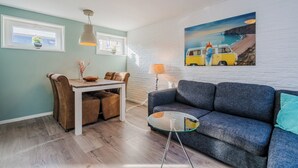 Una sala de estar moderna con un sofá gris, una mesa de comedor con sillas, una planta colgante y un cuadro de una furgoneta en una playa.